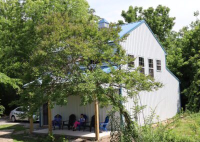 A metal-clad house with a front porch where two people sit, surrounded by lush greenery.